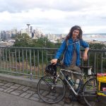 Mikey stands with his bicycle in front of a railing overlooking the Seattle Skyline and the Puget Sound
