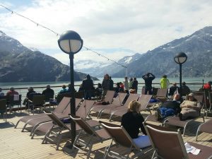 Mikey stands on a cruise ship deck while sailing through Glacier Bay in Alaska. There are stunning snow-capped mountains and glaciers jutting out of the water in the background.