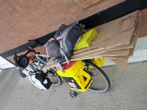 Mikey's fully-loaded bicycle is leaned against a well. Itc arrys 4 panniers, 1 backpack, and a huge cardboard box