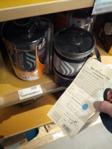 A shelf shows a Jetboil costs $79.95 new. Mikey holds a document titled "REI RETURN TAG" with a prive listed as "$17.85"