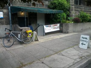 Mikey's bicycle is leaned up against a wall next to a sign that reads "SOUTHEAST PORTLAND TOOL LIBRARY"