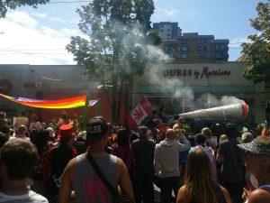 A procession moves down a street full of people. A truck drives down the road with a truck-long sized joint that's emitting smoking from the front. In the back of the truck, a canadian flag with the maple leaf replaced by a cannabis leaf is flying. Behind them, a giant rainbow pride flag serves as a shade cover for the following vehicle.