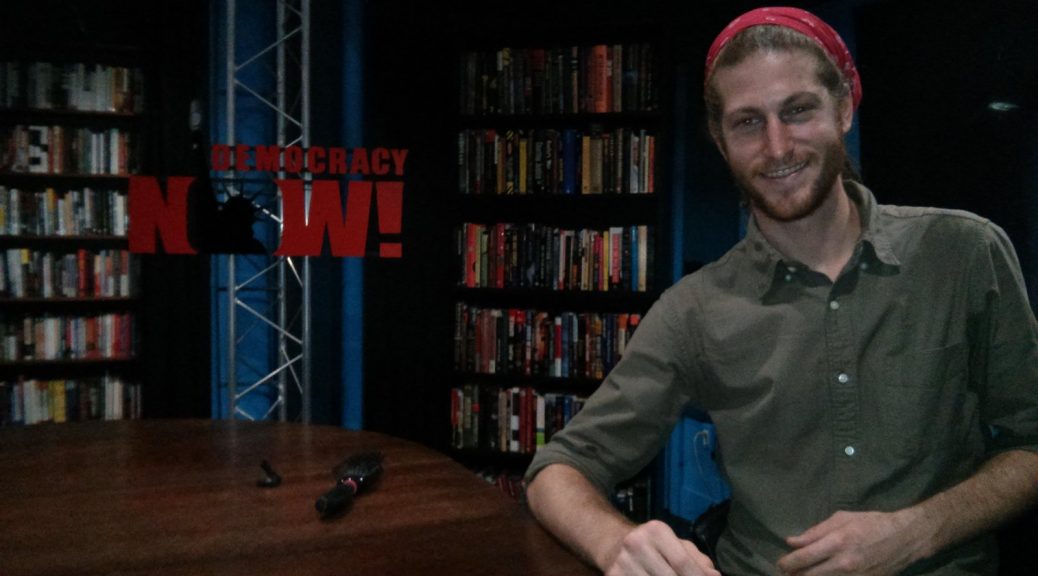 Mikey is sitting at a wooden table, smiling. A sign to his right reads "Democracy Now!" and large bookshelfs sit behind the sign.