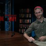 Mikey is sitting at a wooden table, smiling. A sign to his right reads "Democracy Now!" and large bookshelfs sit behind the sign.