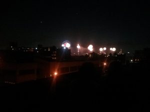 A row of fireworks explode at night with the Empire State Building in the distance.