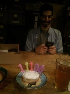 A plate with a sugar-powdered donut has 5 candles in it, labeled "11100". The plate sits next to a tall glass of beer. A box of donuts is behind the plates, next to M smiling on his phone