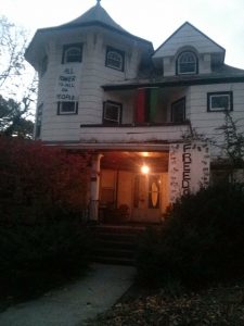 An old, run-down white house has a Panafrican flag hanging from the roof over its second story. A banner hangs from the 3rd story window that reads "ALL POWER TO ALL DA PEOPLE." Another banner hangs on the porch's roof that reads "FREEDOM" with many black handprints on its border.