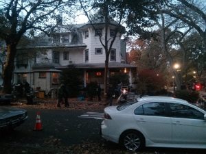 An autumn street is full of leaves and some nearly-bare trees. The front of antique cars can be seen to the left of the frame, and there is an orange traffic cone in the street. Across the road is an old white house. In front of the house are many workers going about various tasks.