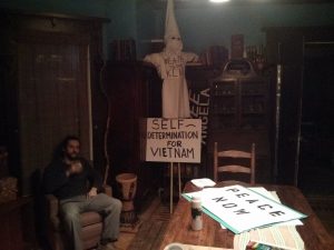 C sits on a chair with a drink in his hand. To his left is an effigy of a KKK member with the words "DEATH TO THE KLAN" written on their chest. A sign under it reads "SELF DETERMINATION FOR VIETNAM" and another behind reads "FREE ANGELA." A sign sitting on the table reads "PEACE NOW."