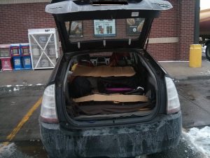A prius from behind with its hatchback open. Inisde is a Brompton bicycle folded and many pieces of cardboard. The black car is dripping with frost and salt. Snow is piled up on the pavement around the car. The car is parked at a gas station.