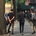 3 old friends walk abrest down a street lined with cars and brownstone steps towards a park.