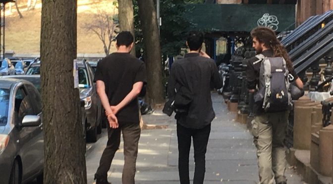 3 old friends walk abrest down a street lined with cars and brownstone steps towards a park.