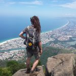 Mikey stands on a rocky cliff overlooking a jungle juxtiposed next to a city with a canal running through it, and emptying-out to the ocean, which stretches out to the horizon.