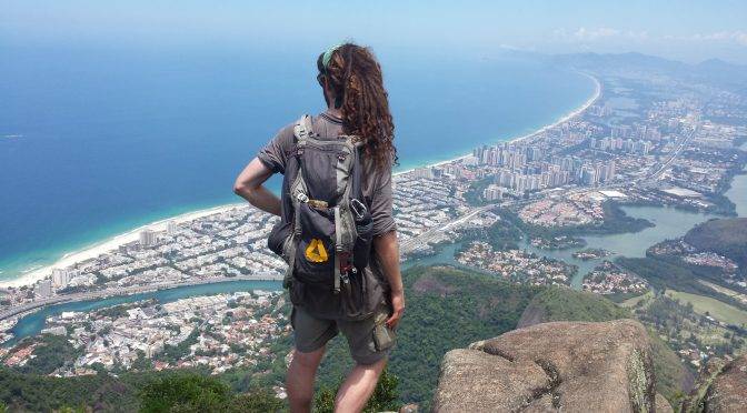 Mikey stands on a rocky cliff overlooking a jungle juxtiposed next to a city with a canal running through it, and emptying-out to the ocean, which stretches out to the horizon.