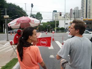 A woman and a man wait in front of a man wearing red holding a red flag that reads "PARE". Across the street there are high rises to the right of the road and large green trees to the left of the road.