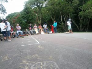 A group of people stand with skateboardsa nd helmets on pavement. Behind them are lush, green trees.
