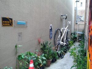 A narrow ally is flanked by many potted plants and a giant PVC bicycle whoose saddle is maybe 7ft high. A sign reads "As Delicias Da Descida." Another sign reads "Bicicletaria"
