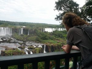 Mikey stands facing away from the camera leaning on a wooden railing. The landscape in front of them is a lush green jungle with a huge river flowing through it. In the background, massive waterfalls gush water into the river.