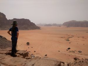 Mikey stands atop a sandstone cliff looking away from the camera, surveying the desert landscape with massive sandstone & granite rocks jutting up out of the sand.