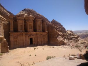 A huge, majestic facade carved out of a sandstone cliff is seen. To the right, the Jordan flag is flapping in the wind.