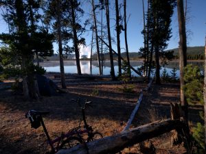 Mikey's bicycle is parked in the woods. Behind it is a tent. Behind that is a large lake. Behind that is an immense tower of steam being produced in the distance