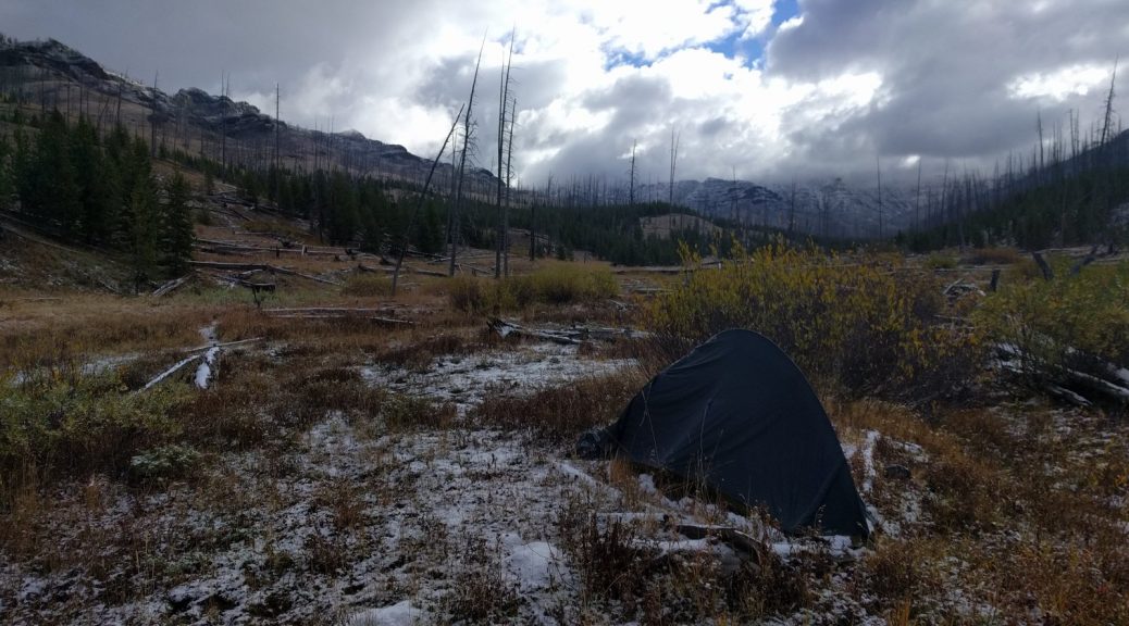 Mikey's tent is setup in a field. There is light snow on the ground and many clouds in the sky. In the distance, snow-capped mountains can be seen.
