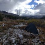 Mikey's tent is setup in a field. There is light snow on the ground and many clouds in the sky. In the distance, snow-capped mountains can be seen.