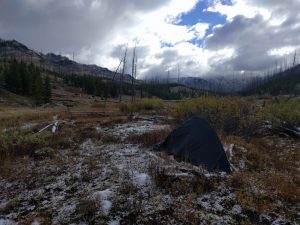 Mikey's tent is setup in a field. There is light snow on the ground and many clouds in the sky. In the distance, snow-capped mountains can be seen.