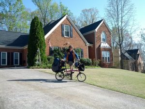 Mikey stands behind his brompton bicycle smiling in front of a brown-bricked suburban home with short-cut, sterile weeds and cement. Towers of trees stand with thin green leaves blooming behind the house.