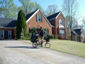 Mikey stands behind his brompton bicycle smiling in front of a brown-bricked suburban home with short-cut, sterile weeds and cement. Towers of trees stand with thin green leaves blooming behind the house.