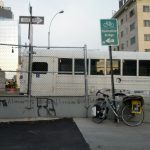 Mikey's bicycle leans up against a sign reading "Manhattan Bridge" with a picture of a bicycle on it. Behind a concrete barrier covered in graffiti, a white bus with dark tinted windows drives past with the inscription "Brooklyn Transportation Corp"