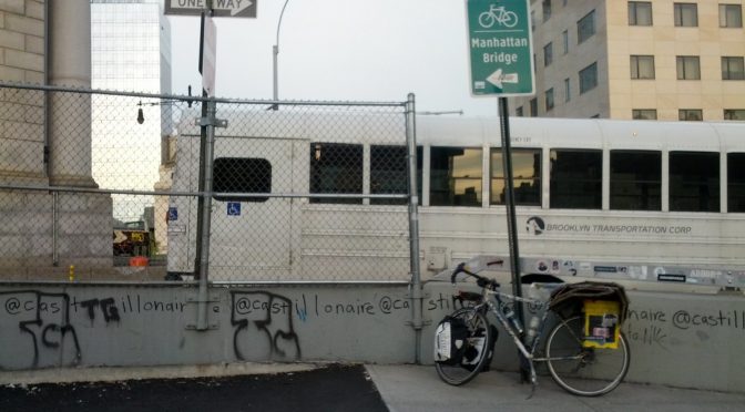 Mikey's bicycle leans up against a sign reading "Manhattan Bridge" with a picture of a bicycle on it. Behind a concrete barrier covered in graffiti, a white bus with dark tinted windows drives past with the inscription "Brooklyn Transportation Corp"