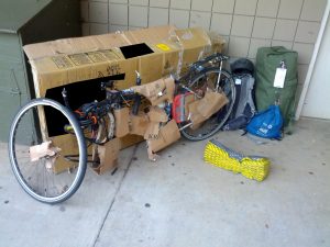 Mikey's bicycle is disassembled, wrapped in cardboard, and just unpacked. Behind it is a huge cardboard bicycle box. Next to it is a backpack, a duffel bag, and a small tote. Mikey's sleeping pad lay crumpled next to his bicycle.