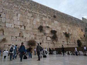 Mikey is walking away from the Western Wall of the Temple Mount in the old city of Jerusalem. They are looking on past the wall that divides the genders, and up to the bridge that climbs to the Temple Mount.