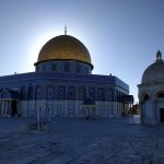 The sun low in the sky, rising in the background. It is near-symmetrically silhouetting the golden top of the Dome of the Rock atop the Temple Mount in the old city of Jerusalem (Al-Quds).