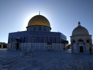 The sun low in the sky, rising in the background. It is near-symmetrically silhouetting the golden top of the Dome of the Rock atop the Temple Mount in the old city of Jerusalem (Al-Quds).