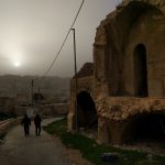 A narrow steet winds through a nearly-abandoned residential neighborhood in Occupied Al-Khalil. A city landscape climbs a hiltop in the distance. Above it, a star tries to shine through a grey fog. Where the road curves to the right, a rock is painted white and blue with a star of David on it.