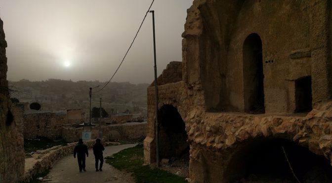 A narrow steet winds through a nearly-abandoned residential neighborhood in Occupied Al-Khalil. A city landscape climbs a hiltop in the distance. Above it, a star tries to shine through a grey fog. Where the road curves to the right, a rock is painted white and blue with a star of David on it.