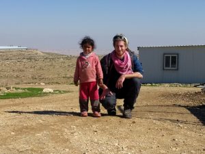 Mikey squats on a dirt road, smiling. Next to him is a young girl, not smiling. Behind them is a small caravan. In the distance, a huge shred of a factory farm can be seen.