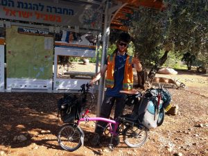 Mikey stands with his bicycle in front of a sign that reads "Welcome to the Northern Portal of the Israel National Trail". Behind the sign is an Olive Tree.