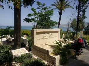 Mikey's bicycle is leaned-up against a grave that reads "משה הס" (Moses Hess). Atop Mikey's saddle sits a red bandanna. The cemetary has many green plants and palm trees.Behind the cemetary is the Sea of Galilee.
