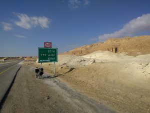 Mikey's bicycle is leaned up against a sign that reads "172" and "Eilat" (in Hebrew, Arabic, and English). The road bends right along impressive canyons and a desert landscape.