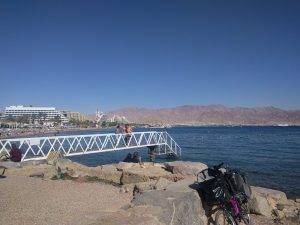 Mikey's bicycle is leaned-up against a rock wall along a beach. The beach's shoreline curves around to Aqaba in the distance. Large hotels and palm trees line the beach.