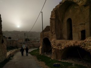 A narrow steet winds through a nearly-abandoned residential neighborhood in Occupied Al-Khalil. A city landscape climbs a hiltop in the distance. Above it, a star tries to shine through a grey fog. Where the road curves to the right, a rock is painted white and blue with a star of David on it.